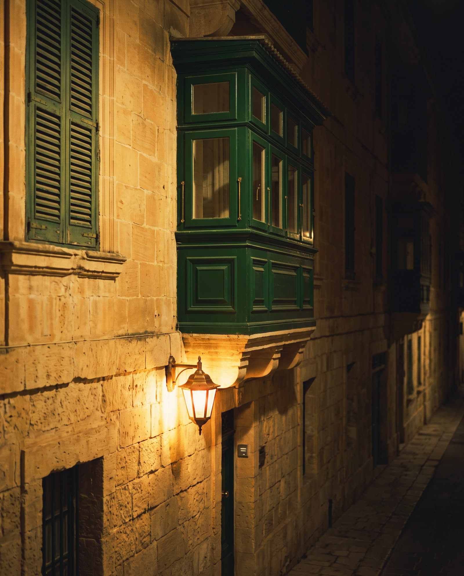 A green Maltese enclosed balcony on a limestone facade at night, a lantern lit below it