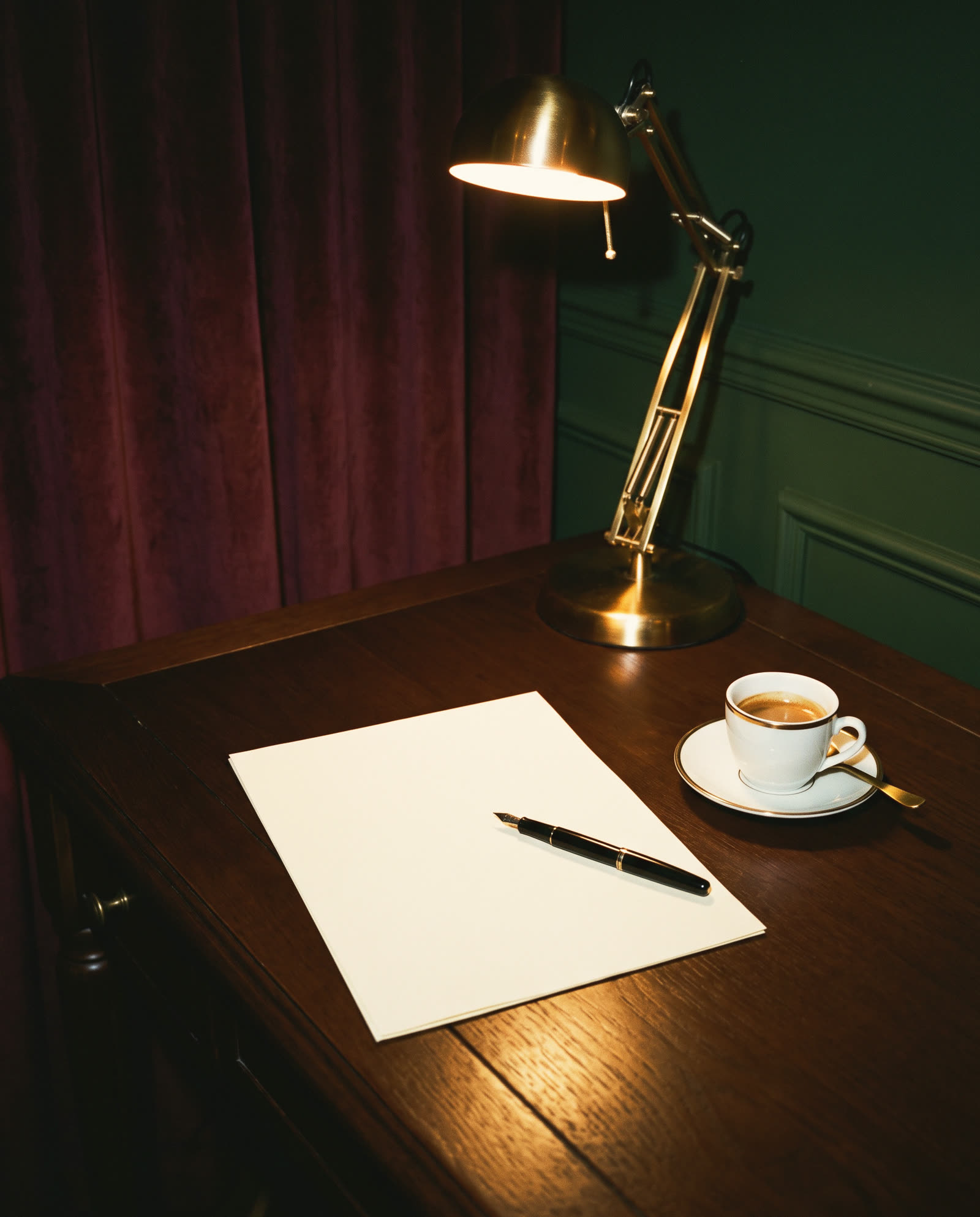 A fountain pen on a blank sheet of cream paper, an espresso and a brass desk lamp