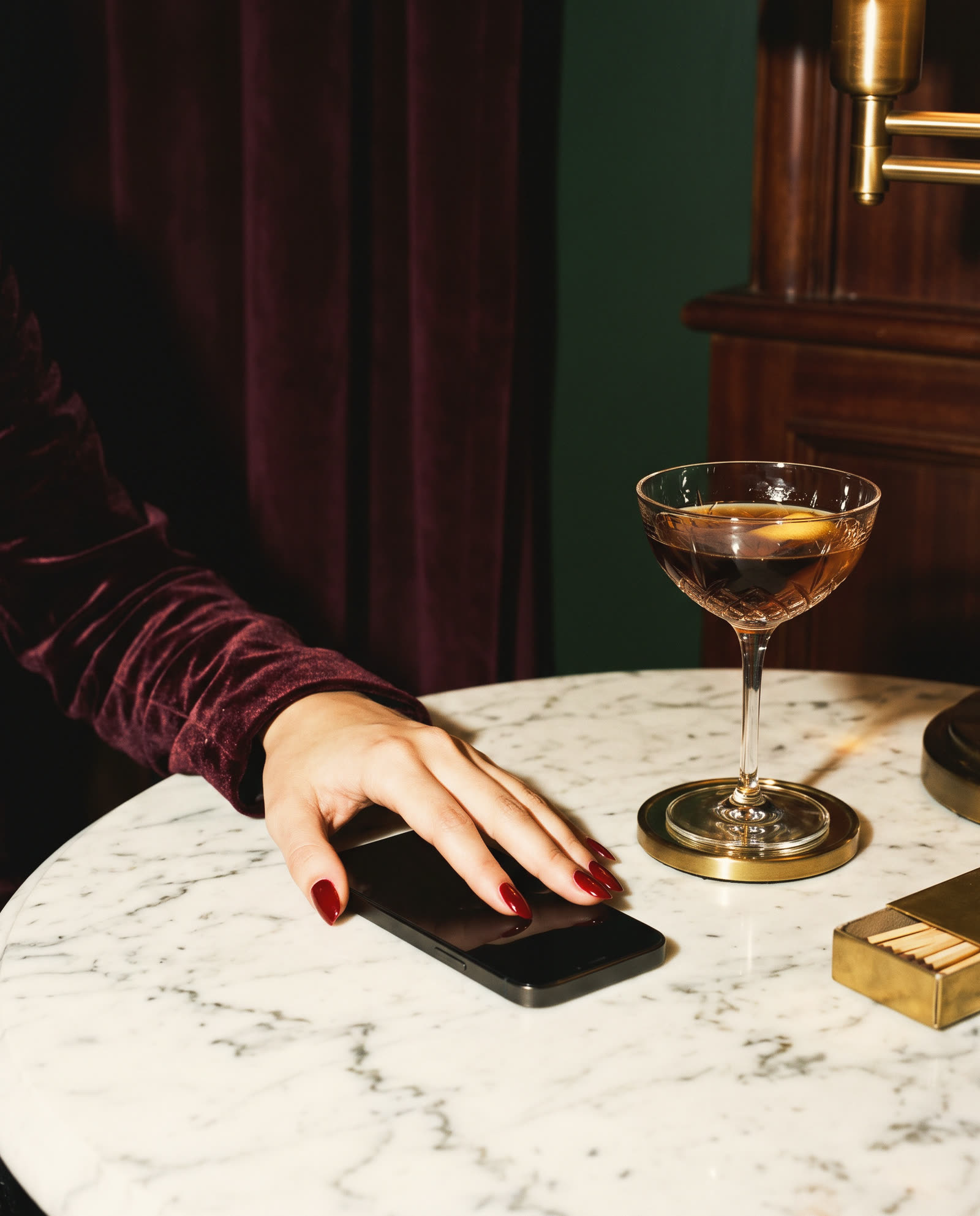 A hand with red nails resting a phone face down on a marble table beside a coupe glass