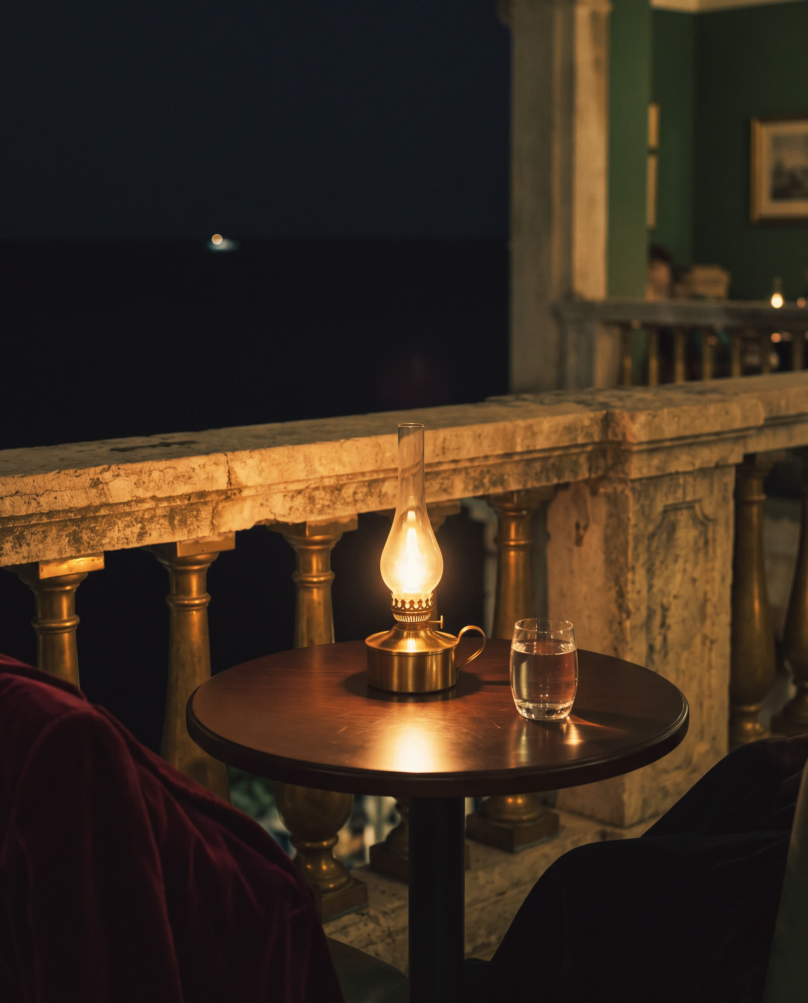 A small terrace table at night with a lit oil lamp and a glass of water, the sea dark beyond the balustrade
